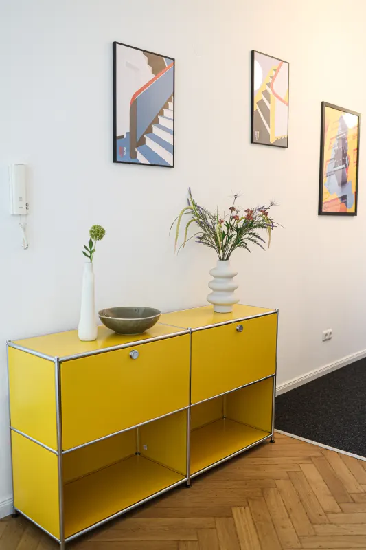 Gelbes USM-Haller-Sideboard mit Vasen vor drei gerahmten Bauhaus-Grafiken auf Fischgrätparkett