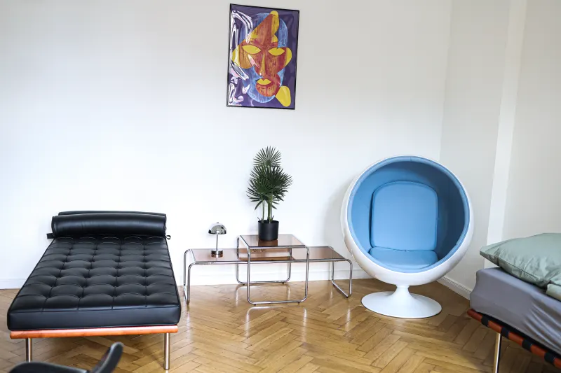 Zweiter Raum mit schwarzem Daybed, blauem Ball Chair und verchromten Beistelltischen auf Parkett